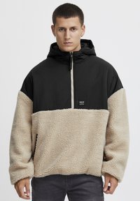 Giacca a pullover con cappuccio nera e beige, caratterizzata da una parte inferiore in pile, una parte superiore liscia, una cerniera frontale e tasche laterali.