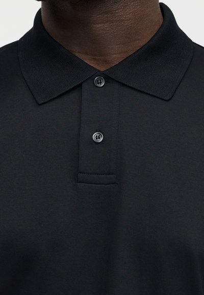 Κοντινό πλάνο ενός ατόμου που φοράει μαύρο πόλο μπλουζάκι με ριμπ κολάρο και δύο μαύρα κουμπιά κλεισμένα στη λαιμόκοψη.