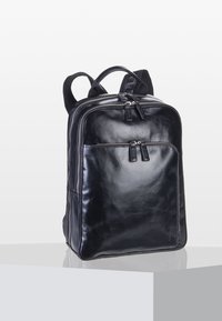 Sac à dos en cuir noir avec compartiments zippés à l'avant et sur le dessus, bretelles rembourrées et une poignée de transport sur le dessus, posé sur une surface réfléchissante.