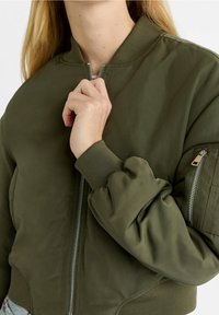 Olijfgroene bomberjack heeft een ritssluiting aan de voorkant, geribde manchetten en een met rits afsluitbare zak op de mouw; gemaakt van glad, lichtgewicht materiaal.