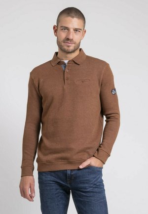 Homme aux cheveux courts portant un polo à manches longues marron et un jean bleu, debout avec une main dans la poche devant un fond uni.