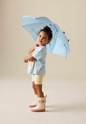 Malé dieťa oblečené v svetlomodrej košeli, žltých krátkych nohaviciach a gumákoch, drží modrý dáždnik s dinosauřími tŕňmi a očami.