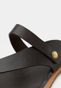 Καφέ δερμάτινο σανδάλι με υφασμένη λουρίδα και διακοσμητικό ορειχάλκινο κουμπί. Η σόλα είναι ανοιχτό καφέ, σε αντίθεση με το σκουρόχρωμο πάνω μέρος. Μινιμαλιστικός σχεδιασμός.