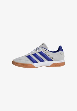 Zapatilla de ante gris con acentos de cuero azul y tres rayas azules. Presenta una suela de goma y "SPEZIALIST" en negro en el costado.