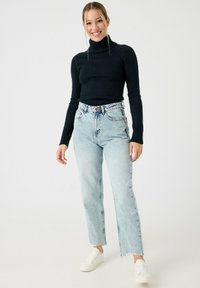 Suéter de punto negro con cuello alto, pantalones vaqueros de cintura alta en un azul claro con dobladillos deshilachados, complementados con zapatillas blancas. Postura erguida con las manos relajadas.