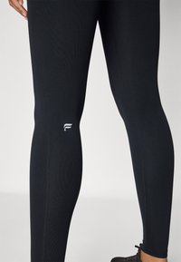 Leggings sportivi neri con un design aderente, texture a costine e un piccolo logo bianco vicino al ginocchio. Leggera lucentezza sul tessuto.