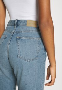 Světle modré džíny s vysokým pasem, ozdobené koženou záplatou v barvě opálené kůže a zadními kapsami, s rovným střihem nohavic a viditelným prošitím.