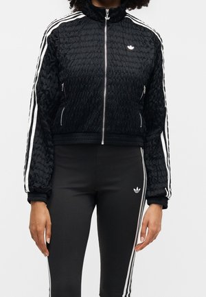 Personne portant une veste de survêtement Adidas noire et des leggings noirs avec des bandes blanches et des logos Adidas sur les manches et la cuisse.