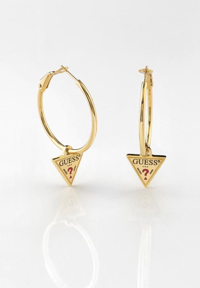 Boucles d'oreilles créoles en or avec pendentifs triangulaires dorés suspendus affichant le logo Guess et un point d'interrogation rouge sur fond blanc.