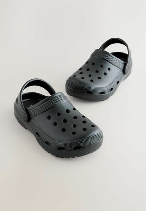 Saboturi negre din plastic cu corp lat, spate deschis și mai multe găuri de ventilație. Talpă texturată cu aderență suplimentară pentru stabilitate.