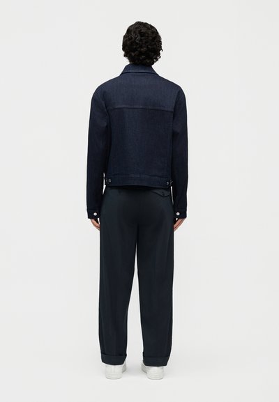 Άτομο με σγουρά μαλλιά, γυρισμένο προς τα πίσω, που φοράει σκούρο μπλε μπουφάν από τζιν, φαρδιά μαύρα παντελόνια και άσπρα παπούτσια σε απλό φόντο.