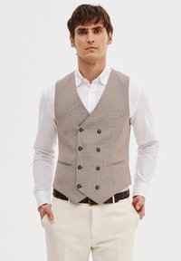 Beżowa kamizelka w wzory z podwójnym zapięciem, z ciemnymi guzikami i dwiema przednimi kieszeniami, noszona na białej koszuli z jasnymi spodniami.