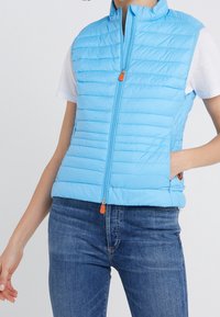 Gilet gonflé bleu clair avec un design matelassé, col montant, fermeture éclair à l'avant avec une tirette orange et poches latérales. Porté par-dessus une chemise blanche.