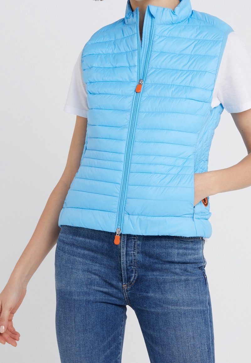 Gilet gonflé bleu clair avec un design matelassé, col montant, fermeture éclair à l'avant avec une tirette orange et poches latérales. Porté par-dessus une chemise blanche.