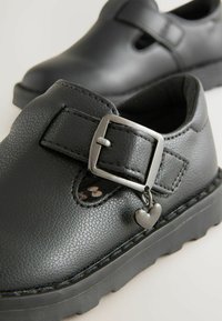 Schwarze Lederschuhe mit runder Zehenform, Schnalle und Herzanhänger. Strukturierte Oberfläche und eine langlebige Gummisohle mit gerilltem Muster.