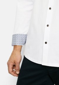 White shirt with a button-down front, featuring a patterned blue cuff with small details. Includes brown buttons and a curved hem.