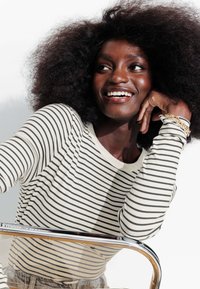 Haut à manches longues rayé en crème et noir. Le mannequin a des cheveux bouclés, souriant, reposant son menton sur sa main. Il comprend un bracelet en chaîne dorée.