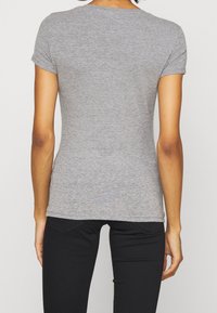 En kvinde iført en tætsiddende grå t-shirt med korte ærmer og sorte bukser, set bagfra mod en ensfarvet baggrund.