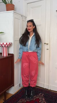 Γυναίκα με μακριά σκούρα μαλλιά φοράει τζιν μπουφάν, κόκκινο παντελόνι με σκίτσα και το κείμενο "AMOR", καθώς και μαύρα μποτάκια, στέκεται σε ένα δωμάτιο κοντά σε μια πόρτα.