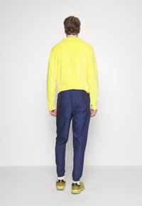 Żółty polarowy sweter z kołnierzem, w połączeniu z granatowymi dresami. Buty są czarne i żółte, z widoczną podeszwą. Prost tło w kolorze białym.