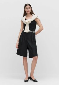 Musta ilmanhihainen paita, muotoillut mustat shortsit ja kerma valkoinen neule, joka on sidottu hartioiden ympärille; kapea malli; tyylikkäät mustat kengät.