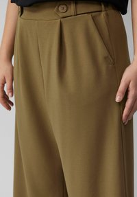 Pantalon vert olive en tissu doux et texturé, avec une fermeture à un bouton et des poches latérales ; il présente un design à plis sur le devant.