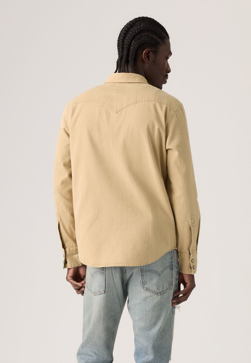 Homme aux cheveux tressés portant une chemise beige à manches longues et un jean bleu clair, de dos sur un fond clair uni.