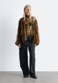 Braune Spitzentbluse mit langen, puffigen Ärmeln und hohem Kragen, über einem schwarzen Oberteil getragen; kombiniert mit locker sitzenden schwarzen Jeans und schwarzen Schuhen.