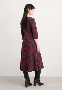 Femeie cu păr lung și închis la culoare, îmbrăcată într-o rochie midi cu flori roșii și bleumarin și cizme cu toc înalte, negre, stând de profil pe un fundal simplu.