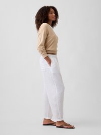 Femme aux cheveux bouclés, vêtue d'un pull beige, d'un pantalon blanc ample et de sandales foncées, sur un fond uni gris clair.