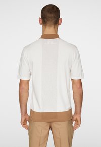 Polo shirt bianco a maniche corte con colletto e orlo marroni. Presenta un motivo a righe verticali testurizzate sul retro. Tessuto in misto cotone.