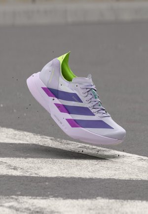 Chaussure de sport gris clair avec des bandes violettes et un intérieur vert, prise en plein vol au-dessus d'un bitume avec des marquages routiers blancs.