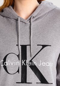 Sudadera gris de tejido suave con un gran logo negro "CK" y el texto "Calvin Klein Jeans" debajo en blanco. Incluye cordones.