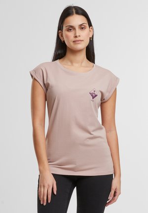 T-shirt mauve clair en tissu doux avec des manches courtes roulées, ornée d'un petit design graphique violet sur la poitrine, associée à un jean noir.