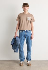 T-shirt beige à manches courtes avec détail de poche, jeans en denim bleu, tenant une veste en denim, baskets marron et blanches, debout contre un fond uni.