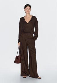 Bruine gebreide trui met V-hals, gecombineerd met hoge wijdvallende broek in een donkerdere bruintint. Houdt een kleine bruine handtas vast en staat tegen een effen achtergrond.