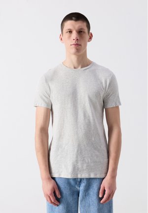 Noor mees seisab, kandes helehallist lühikese varrukaga t-särki ja siniseid teksapükse, taustal tavaline valge sein.