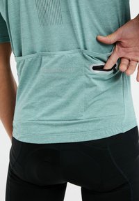 T-shirt de sport vert clair, respirant, avec un motif texturé, poche arrière accessible par une zip, associé à un pantalon noir ajusté.