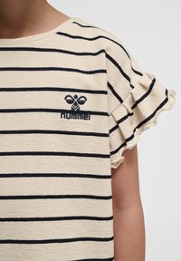 Beige stribet skjorte med marineblå vandrette linjer, med voluminøse ærmer og et broderet logo. Blød tekstur og afslappet design.