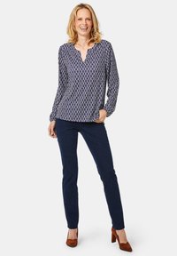 Langarmige marineblauwe blouse met wit geometrisch patroon, V-hals, gecombineerd met donkerblauwe slim-fit jeans en bruine suède hoge hakken.