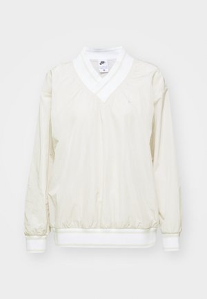 Svetlo bež Nike pulover z V-izrezom, dolgimi rokavi in elastičnimi manšetami. Ima bel pas in majhen vezen logotip na prsih.