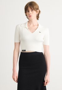 Weißes, geripptes Crop-Top mit V-Ausschnitt und kurzen Ärmeln. Mit einem kontrastierenden grünen Logo auf der Brust. Kombiniert mit einem schwarzen, gerippten Rock.
