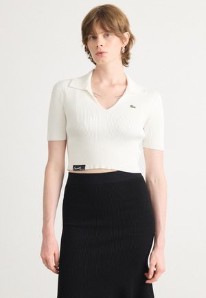 Hvit ribbet crop top med V-hals og korte ermer. Har en kontrasterende grønn logo på brystet. Kombinert med et svart ribbet skjørt.