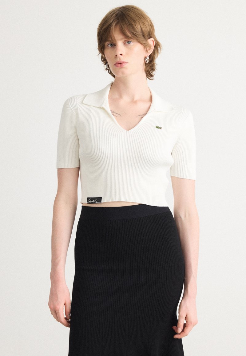 Weißes, geripptes Crop-Top mit V-Ausschnitt und kurzen Ärmeln. Mit einem kontrastierenden grünen Logo auf der Brust. Kombiniert mit einem schwarzen, gerippten Rock.