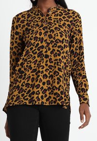 Blusa con stampa leopardata in senape con macchie nere e borgogna, dotata di una chiusura con bottoni e maniche lunghe. Tessuto morbido, vestibilità comoda.
