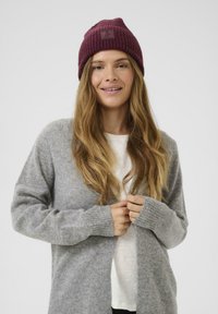 Cardigan grigio con polsini a costine, indossato sopra una camicia bianca. Un berretto di maglia color bordeaux con una toppa quadrata completa l'outfit. Texture morbida visibile.