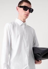 Hvit langermet skjorte med spiss krage, med knappdetaljer. Modellen holder en svart lærveske mens hun har på seg svarte rektangulære solbriller.
