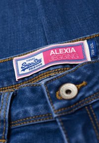 Jeggings de mezclilla en azul oscuro con una etiqueta rosa y blanca que presenta "SuperDry" y "Alexia Jegging". Detalles de costura dorada y botón de metal.
