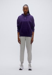 Μωβ φούτερ με κουκούλα, μπροστινή τσέπη και λογότυπο, σε συνδυασμό με γκρι φόρμα. Το μοντέλο φοράει ανοιχτόχρωμα παπούτσια με λεπτομέρειες, στέκεται μπροστά σε απλό φόντο.