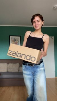 Brown Zalando box with black logo, held by a person in a black tank top and light blue jeans, in a room with green walls.